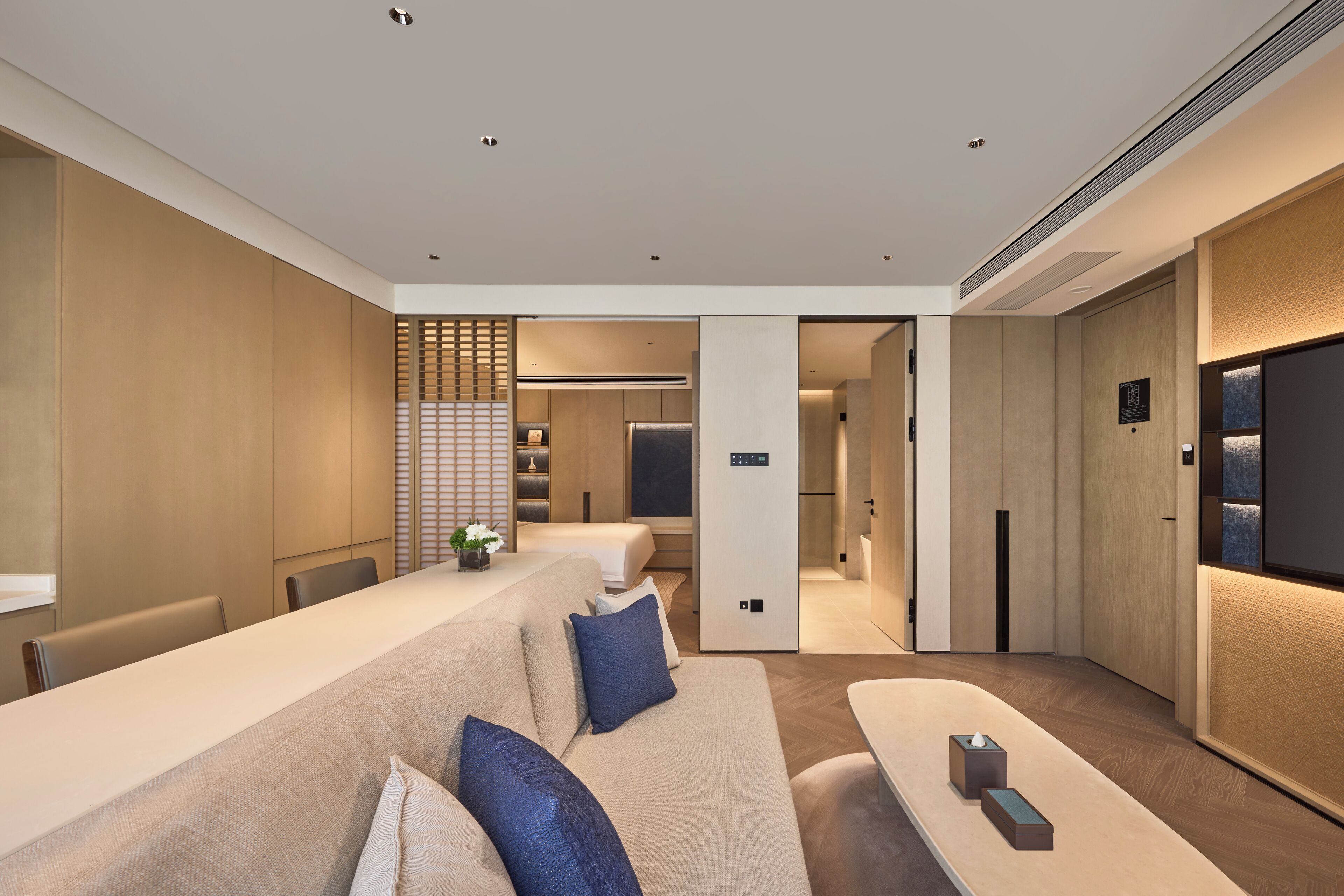 Een moderne hotelkamer met een lange bank, een klein tafeltje en een televisie. Achtergrond is een keukenhoek.