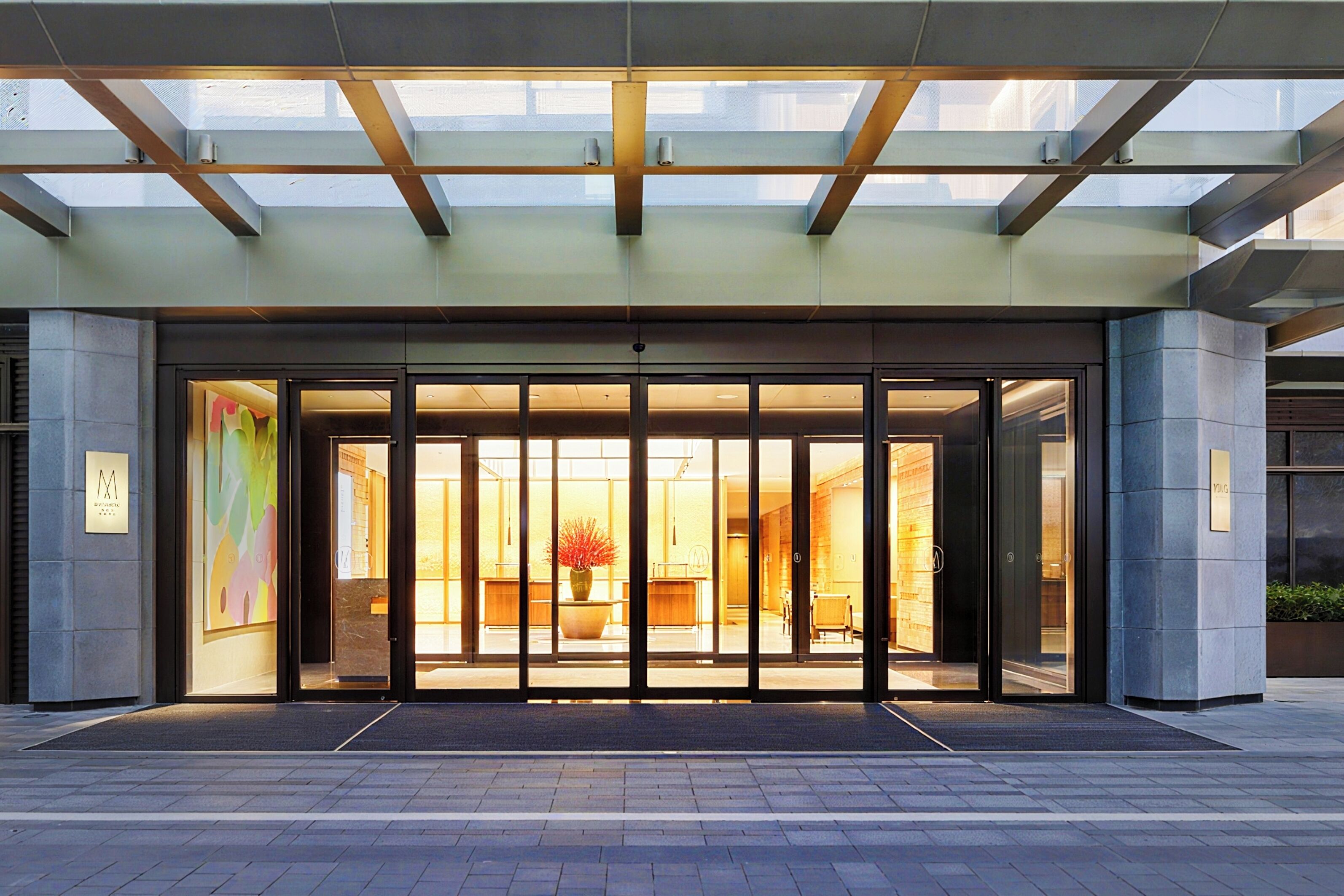 Entrada de hotel con puerta de cristal, interior visible con mostrador de recepción y una planta en maceta.