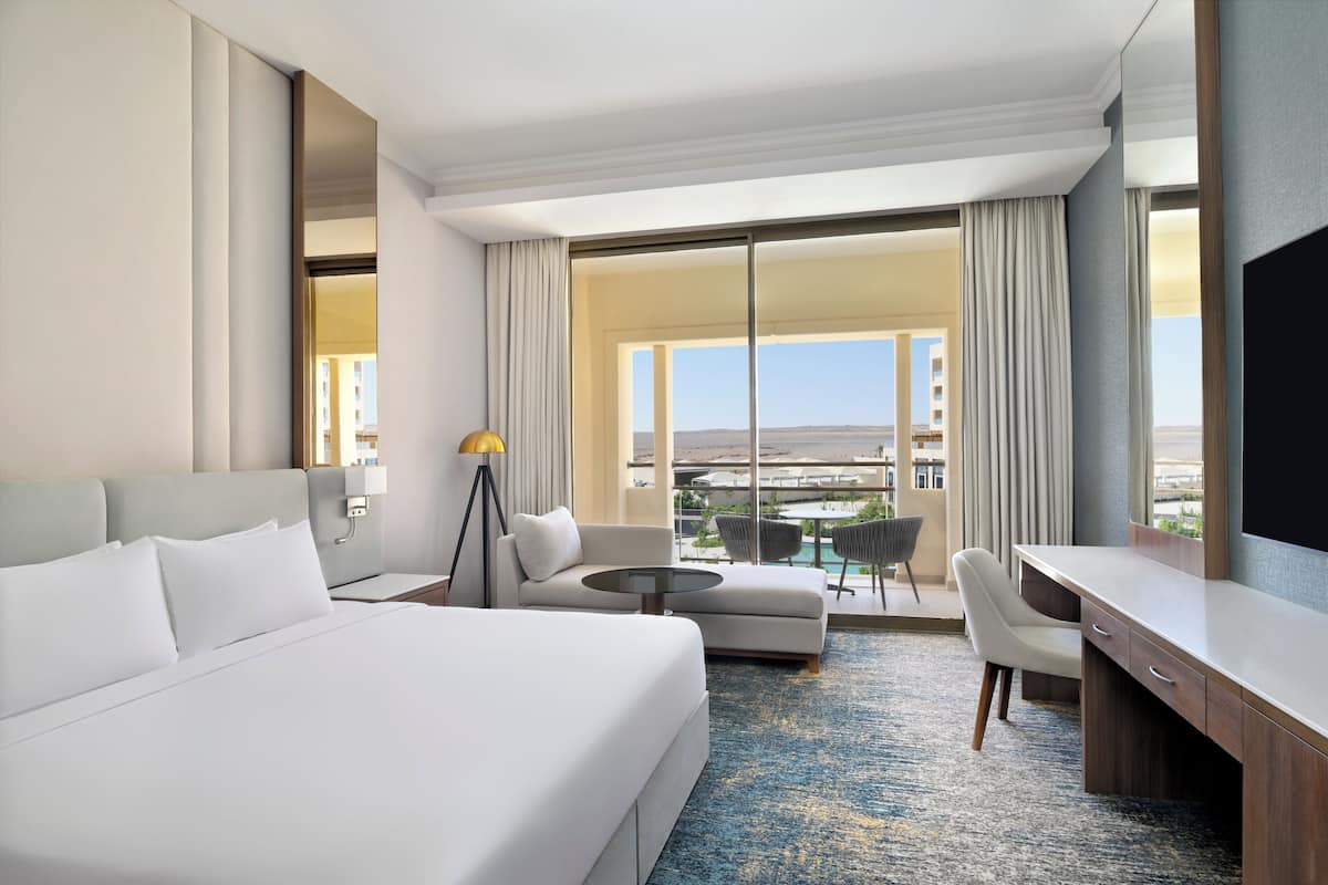 Ein Hotelzimmer mit einem großen Bett, einem Schreibtisch, einem Sessel, einem kleinen runden Tisch und Blick auf die Stadt durch große Fenster.