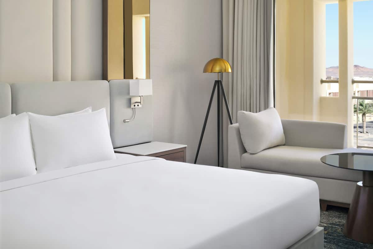 Ein Hotelzimmer mit einem großen Bett, einer Couch, einem kleinen Tisch, einer Lampe, einem Spiegel und Blick ins Freie.