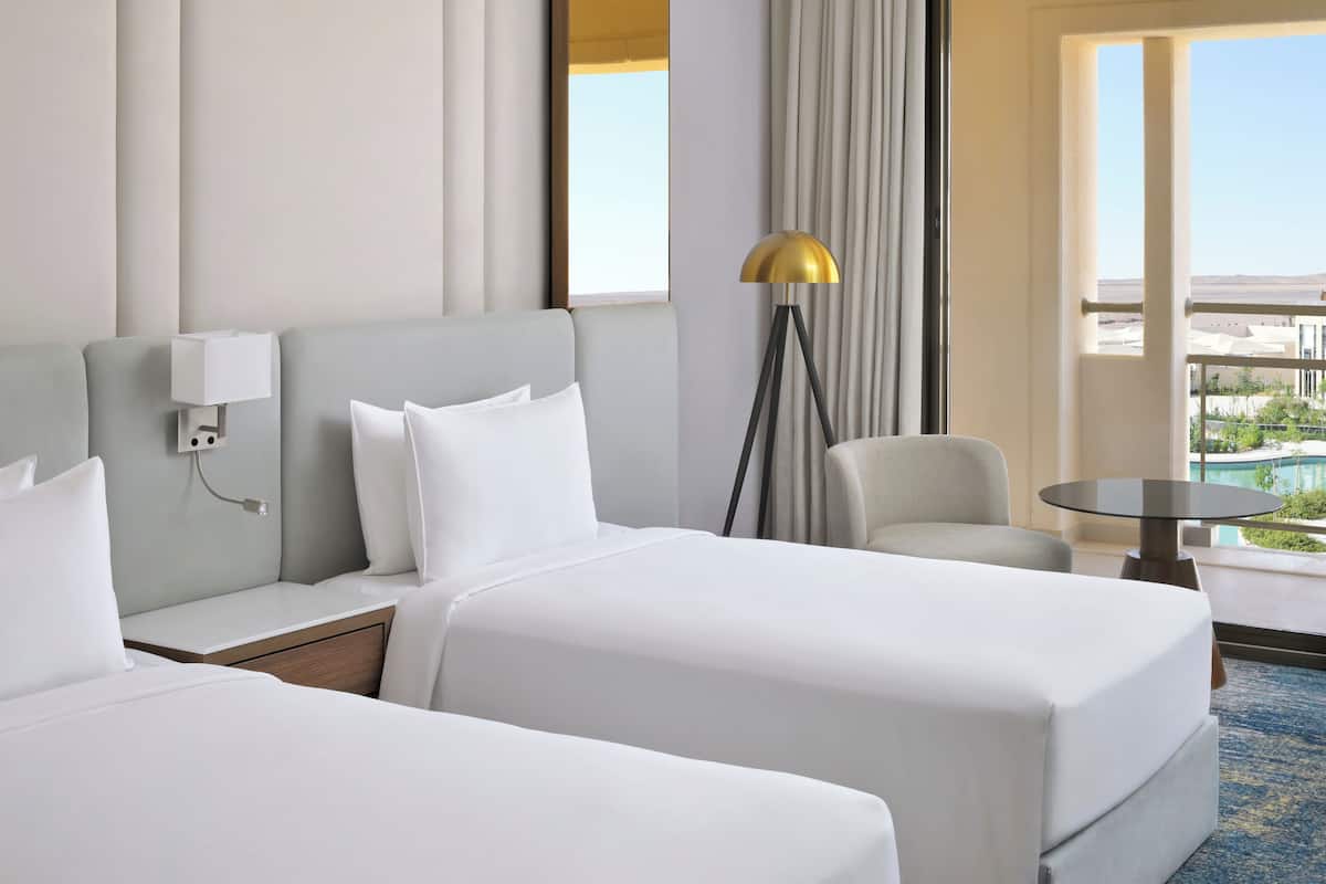 Ein Hotelzimmer mit zwei Betten, einem Nachttisch mit Lampe, einem Sessel, einem runden Tisch und Blick auf einen Pool und die Landschaft.
