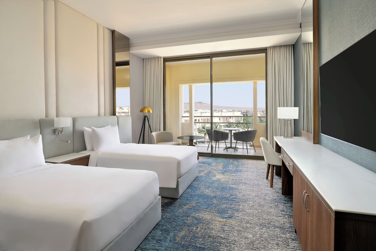 Ein Hotelzimmer mit zwei Betten, einem Schreibtisch, einem Stuhl, einer Lampe und Blick auf die Stadt.
