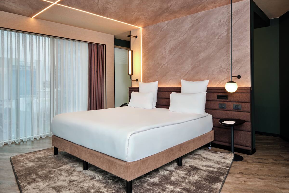 Una habitación de hotel moderna con una cama grande, una mesita de noche, una lámpara de pared y vistas de la ciudad a través de amplias ventanas.