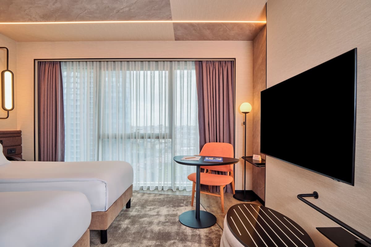 Habitación de hotel con cama, escritorio, silla, televisor y un amplio ventanal con cortinas.