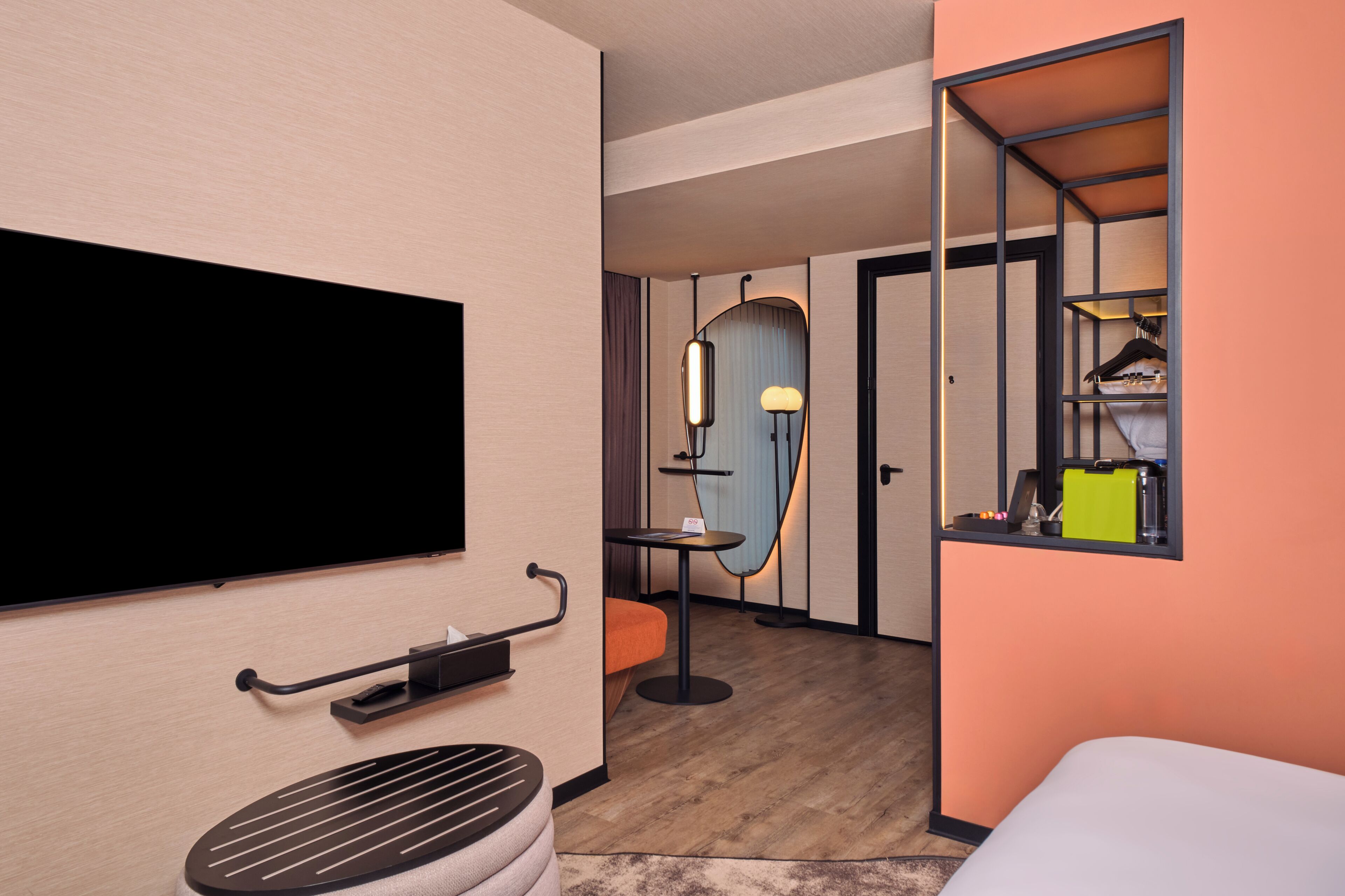 A hotel room with a flat-screen TV, a small round table, a chair, a mirror, and a wardrobe with a hanging rack.