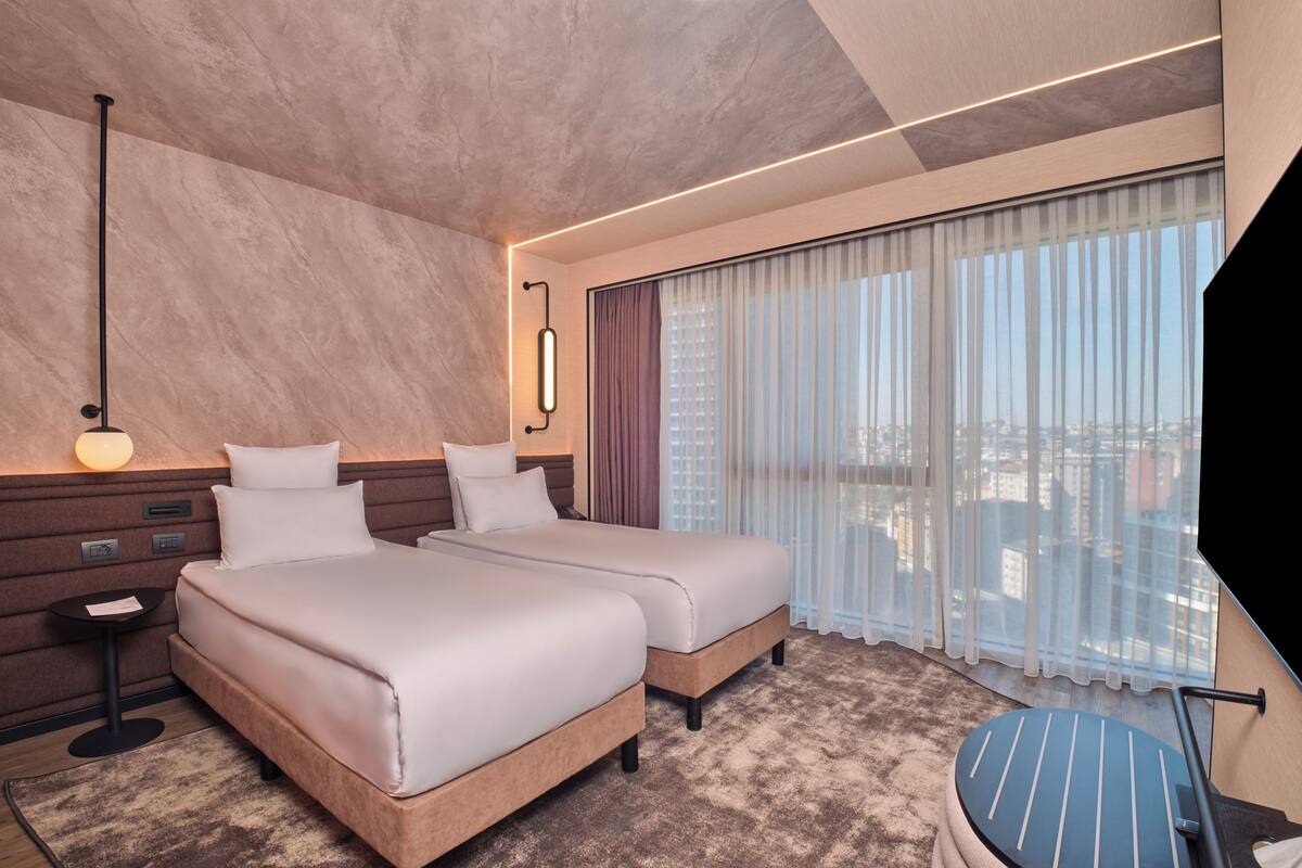 Habitación de hotel con dos camas, un escritorio, una silla, una lámpara y un ventanal grande con cortinas.