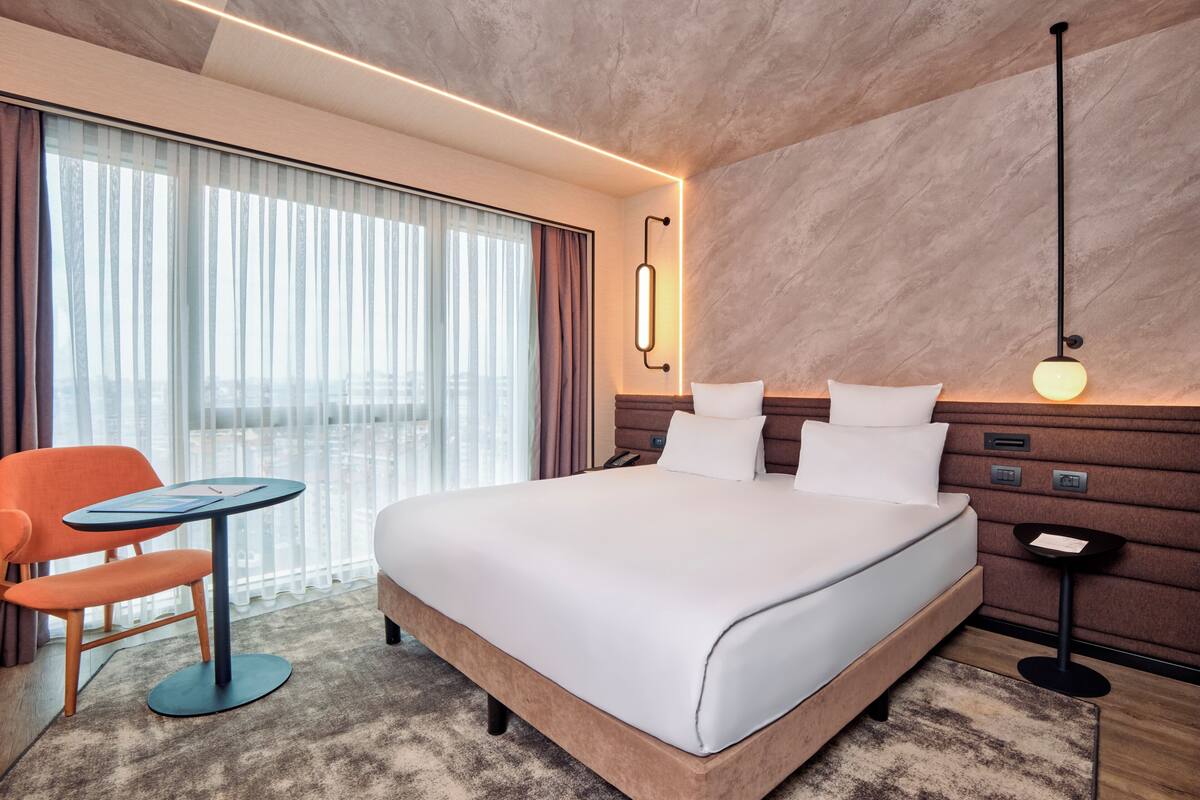 Habitación de hotel moderna con una cama grande, una mesita, una silla y vistas de la ciudad a través de amplias ventanas.
