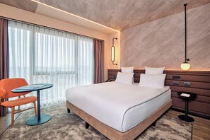 Une chambre dâhĂŽtel moderne, dotĂ©e dâun grand lit, dâune petite table, dâune chaise et offrant une vue sur la ville grĂące Ă de grandes fenĂȘtres.