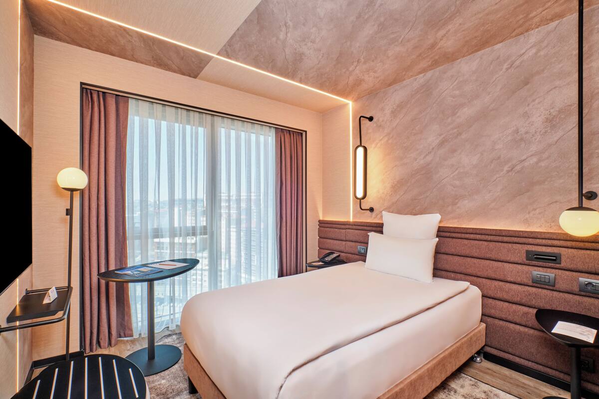Una habitación de hotel con cama, escritorio, silla, televisor y ventana con cortinas.