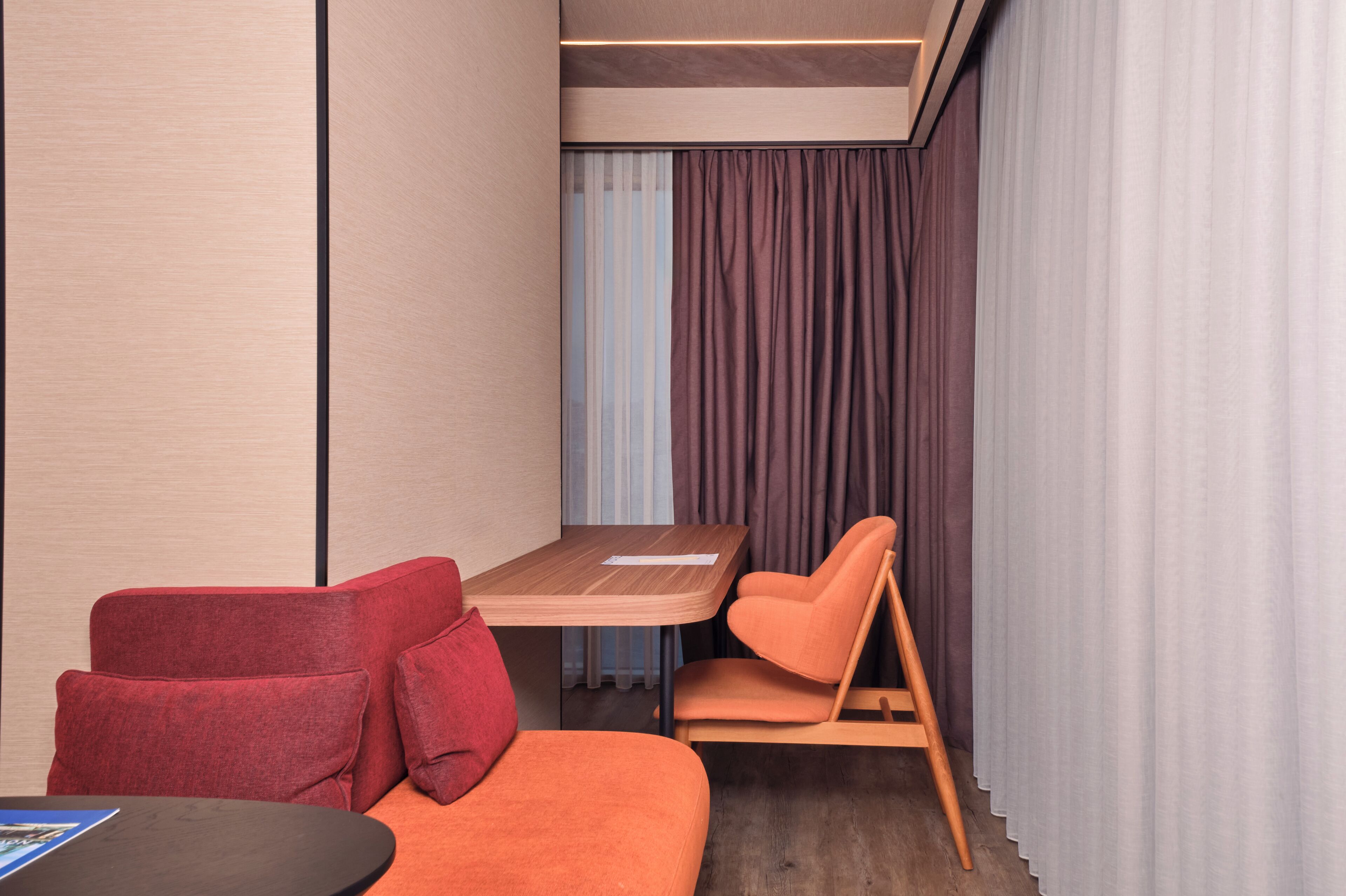 A modern room with a wooden table, orange seating, and a view of the outside through curtains.