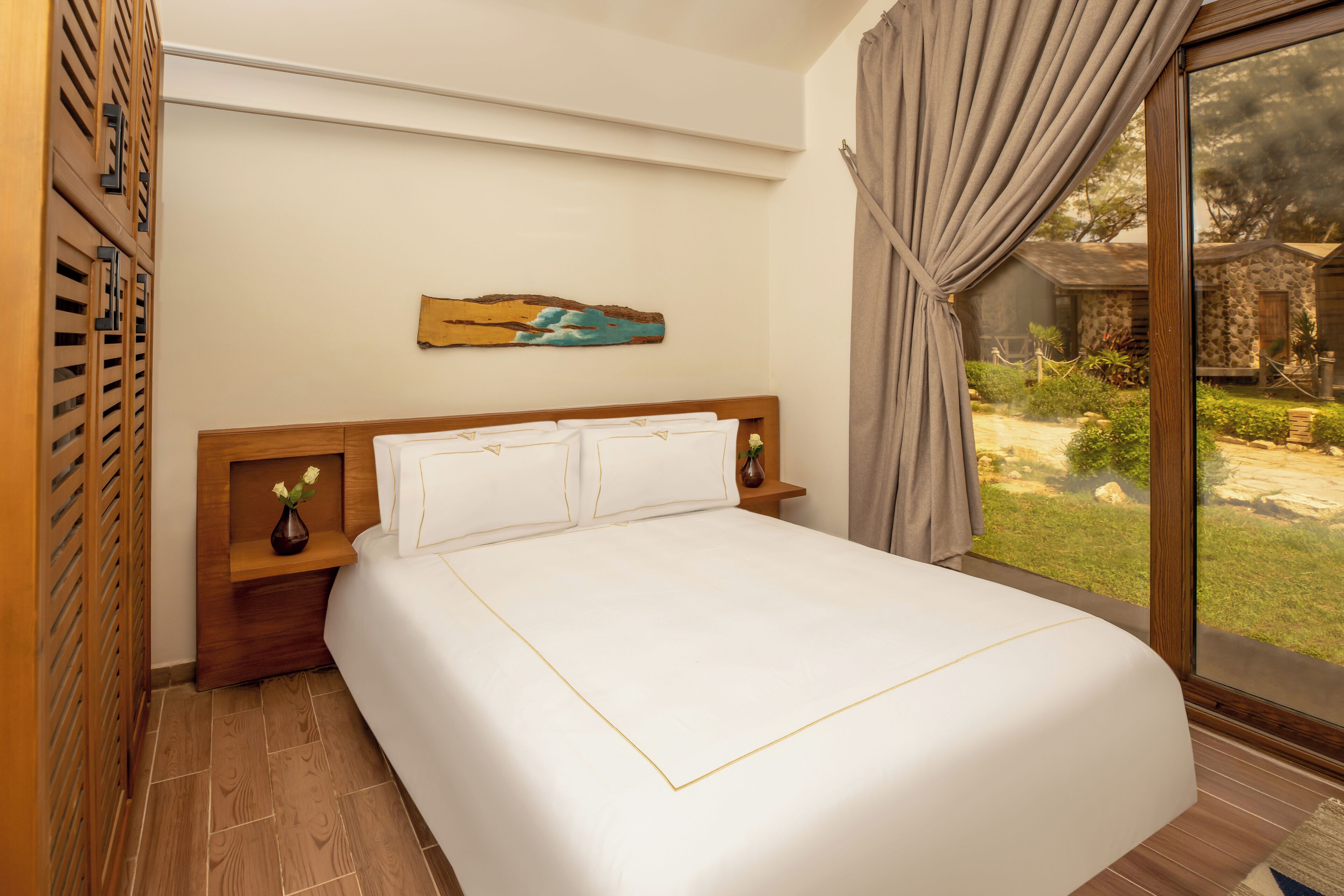 A neatly made bed with white linens and gold trim, a wooden headboard, a nightstand with a vase of flowers, and a painting of a landscape on the wall.