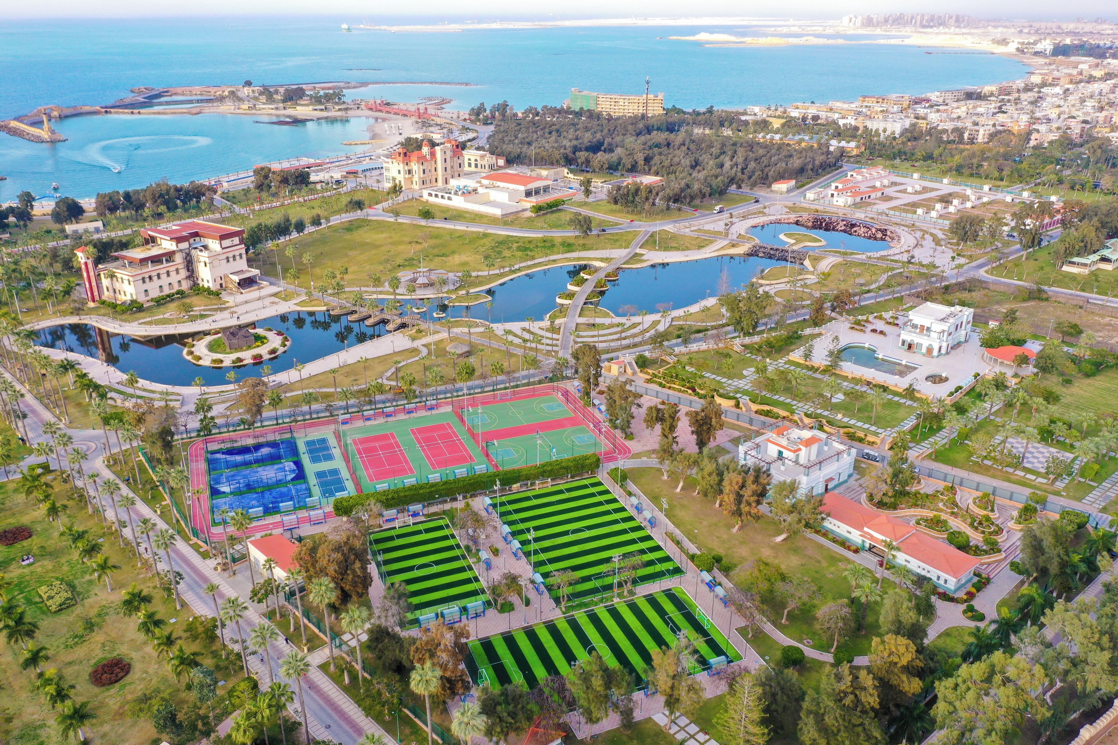 An aerial view of a coastal area with a large sports complex, multiple tennis courts, and a central water feature.