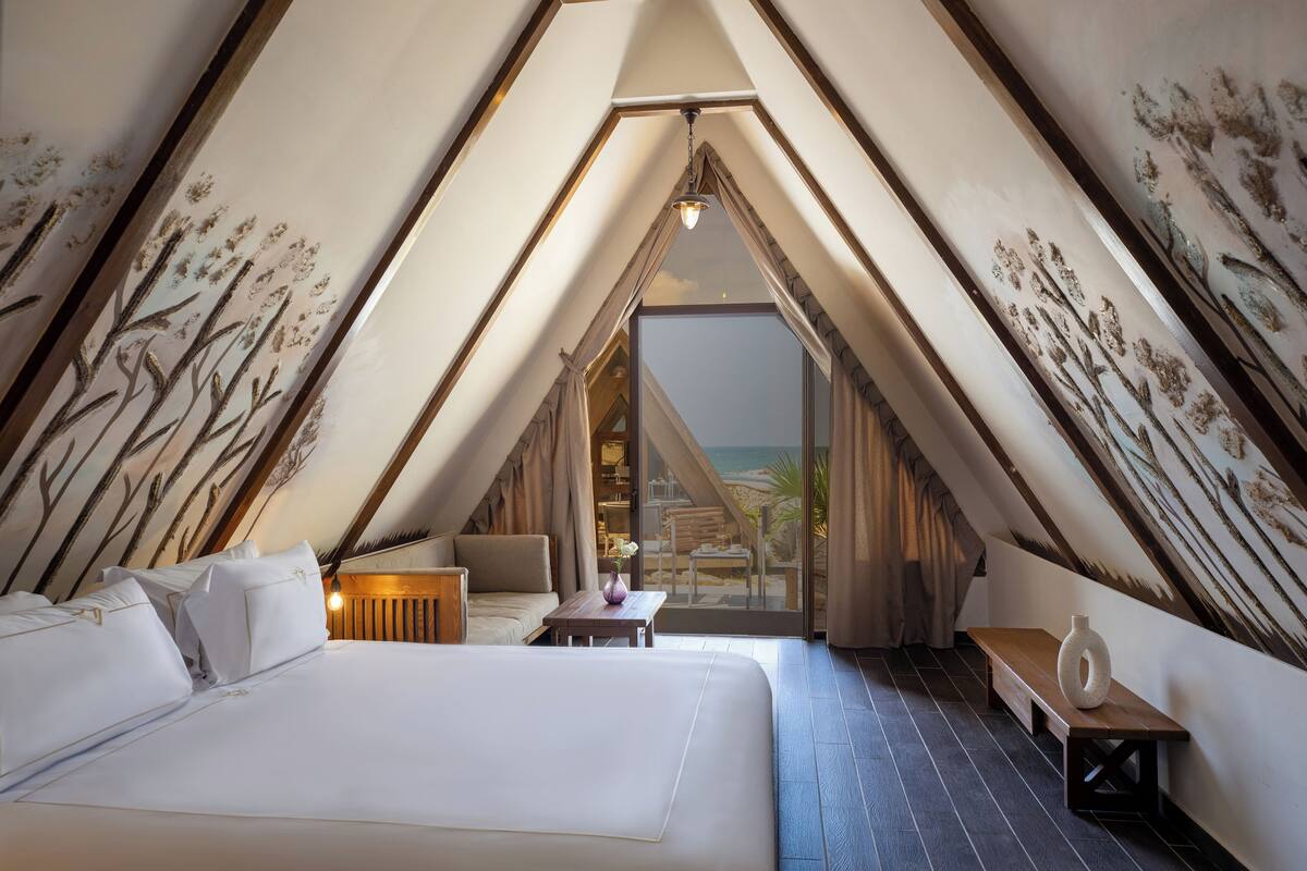 A bedroom with a large bed, a small table, and a bench. There is a view of the beach through a large window.