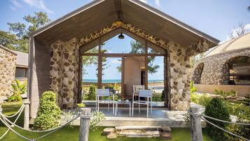 A stone building with a sloped roof and large windows overlooking a beach. There are two white chairs and a table on the patio.
