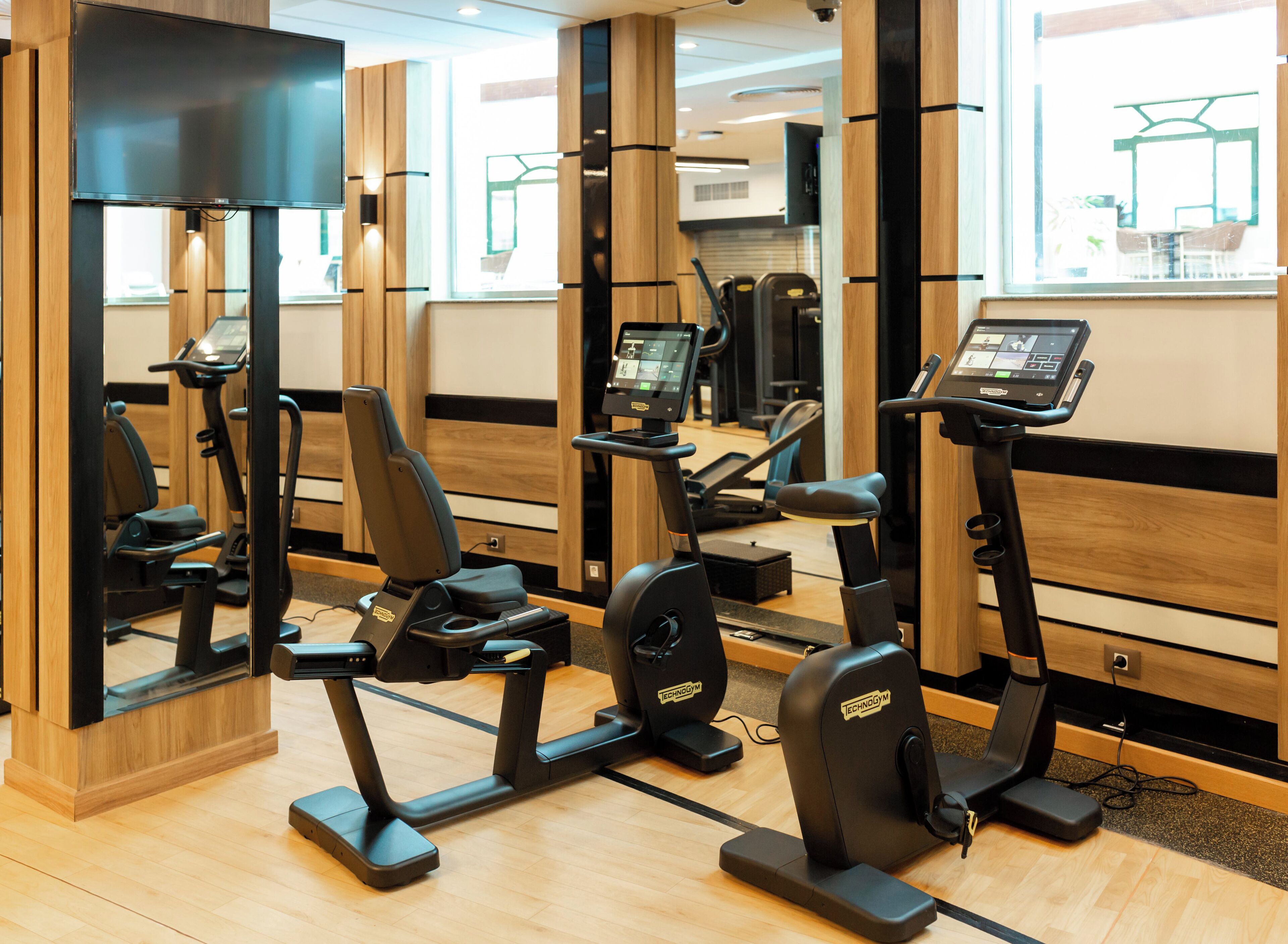 A gym with multiple exercise machines, including treadmills and stationary bikes, arranged in a spacious room with wooden accents and large windows.