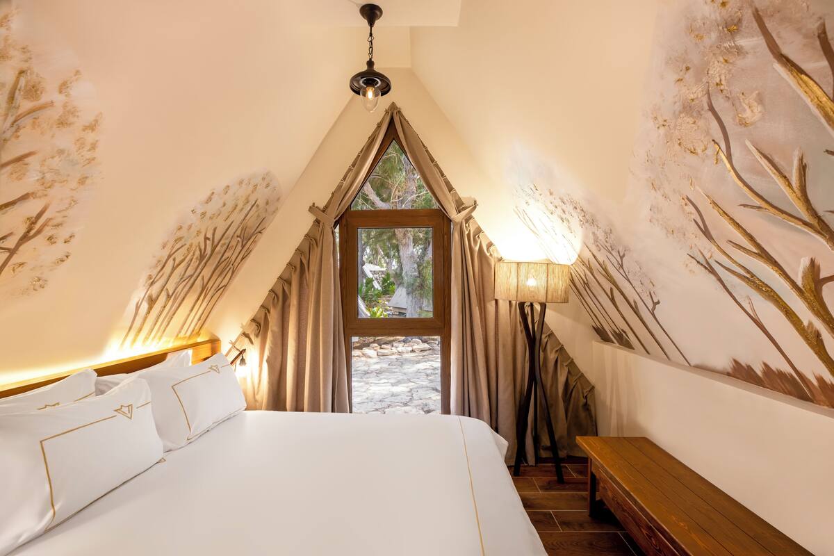 A bedroom with a large bed, a wooden bench, a lamp, and a view of the outdoors through a window.