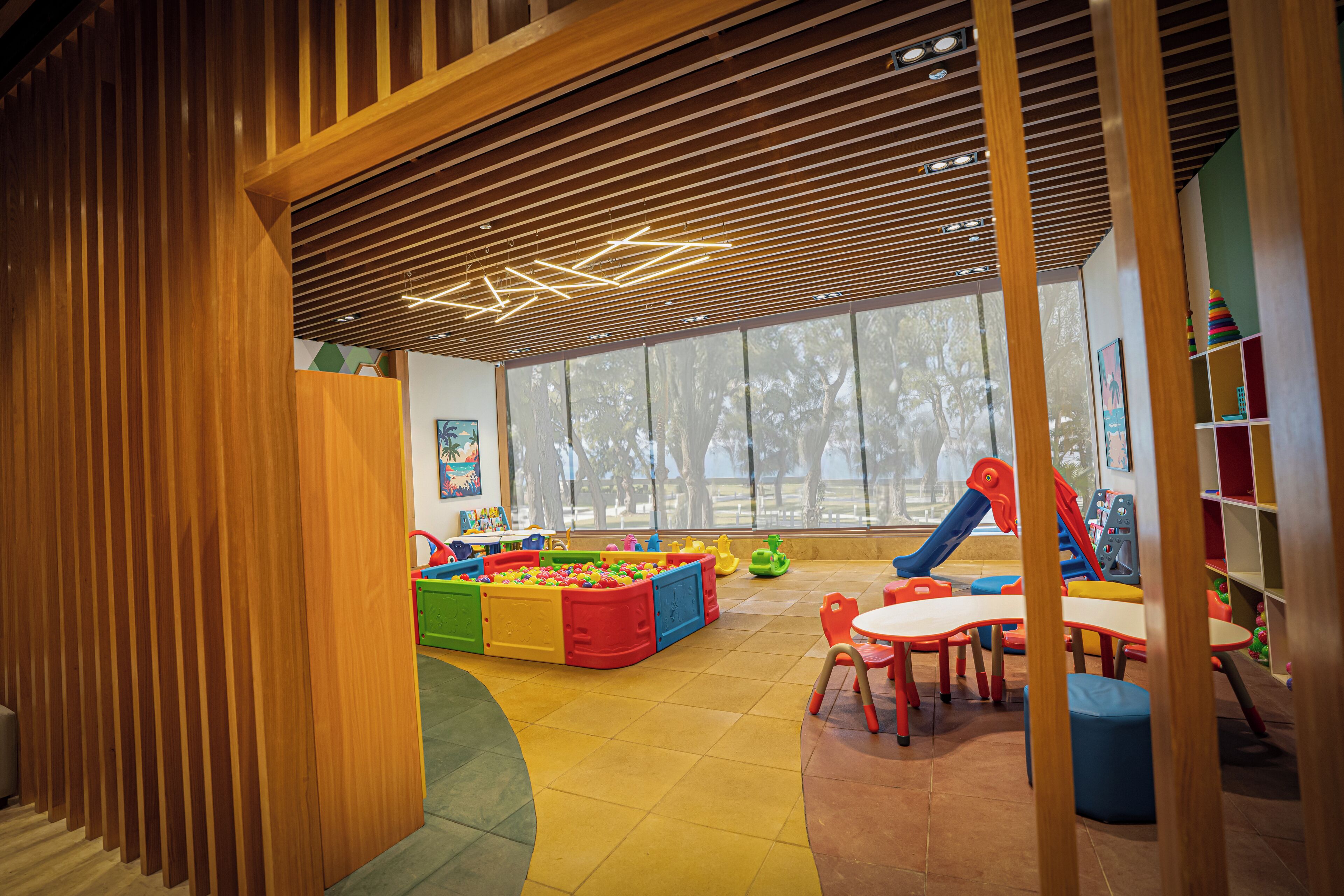 A children's play area with colorful play structures, a large window, and a wooden slatted ceiling.