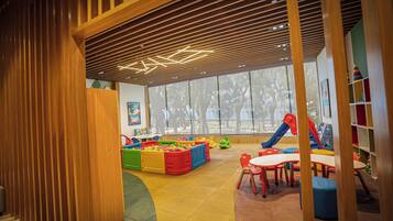 A children's play area with colorful play structures, a large window, and a wooden slatted ceiling.