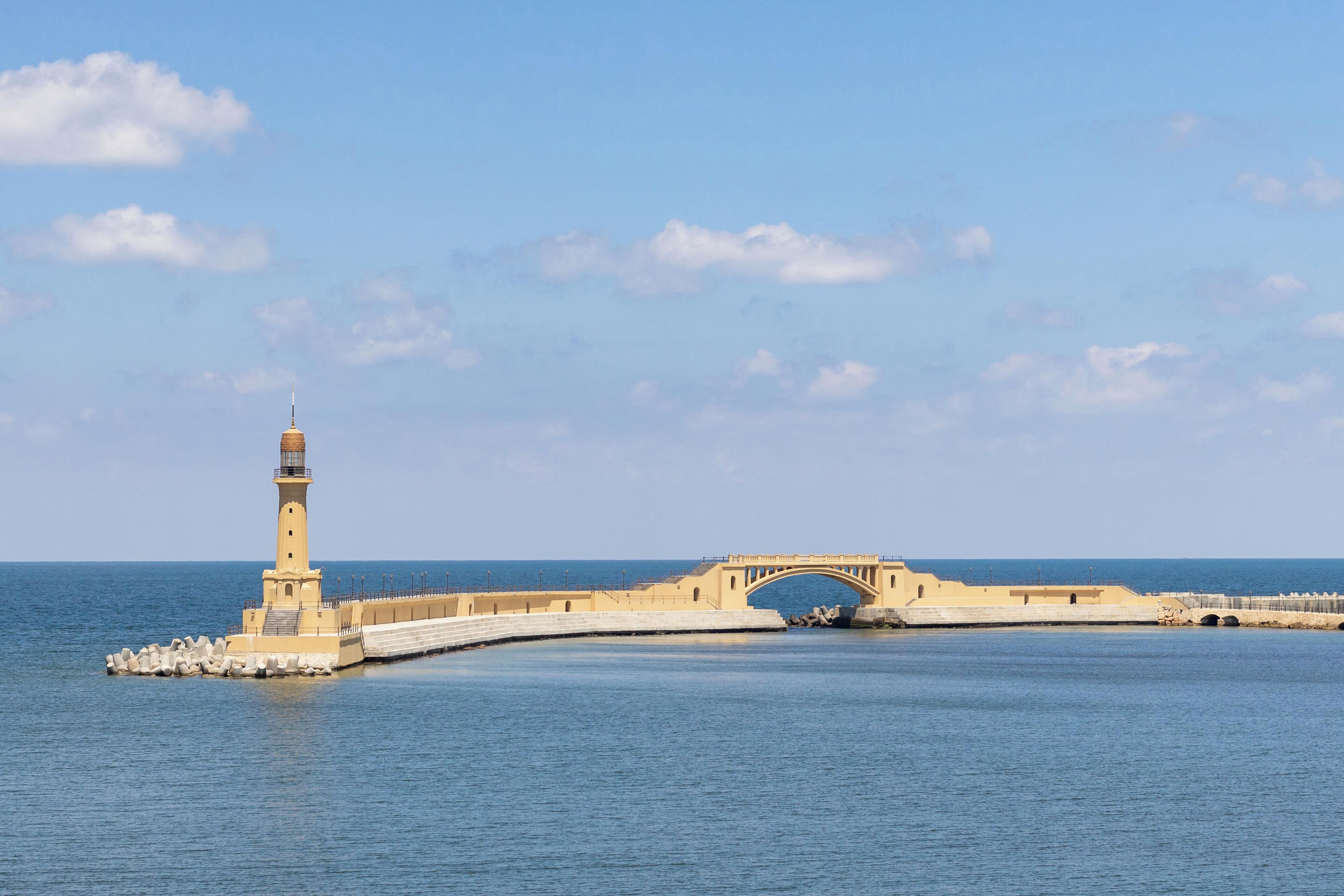 Un puente costero con un faro a la izquierda, que une dos extensiones de tierra sobre aguas azules y tranquilas.