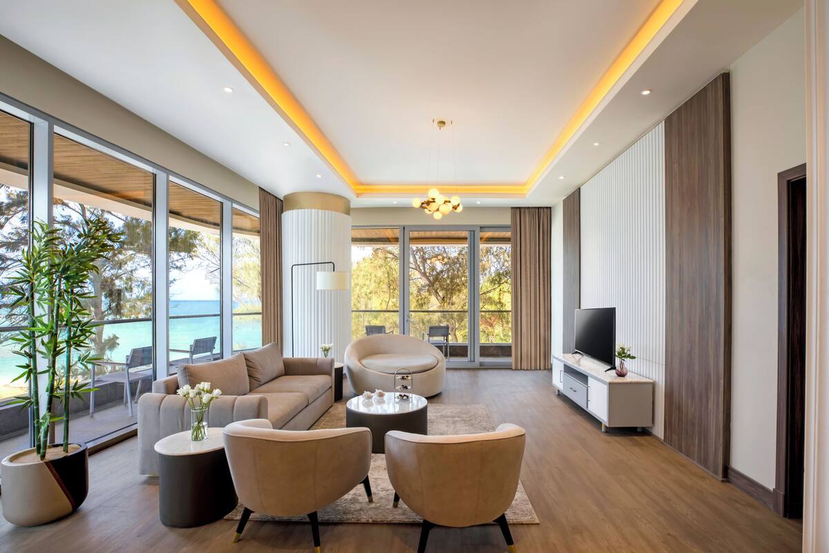 A modern living room with a sofa, armchairs, a coffee table, and a TV. There is a large window with a view of the outdoors.