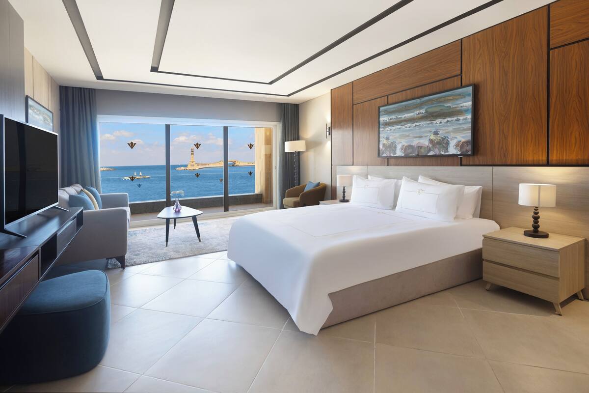 A modern hotel room with a large bed, a TV, a seating area with a view of the sea, and a wooden panel wall.