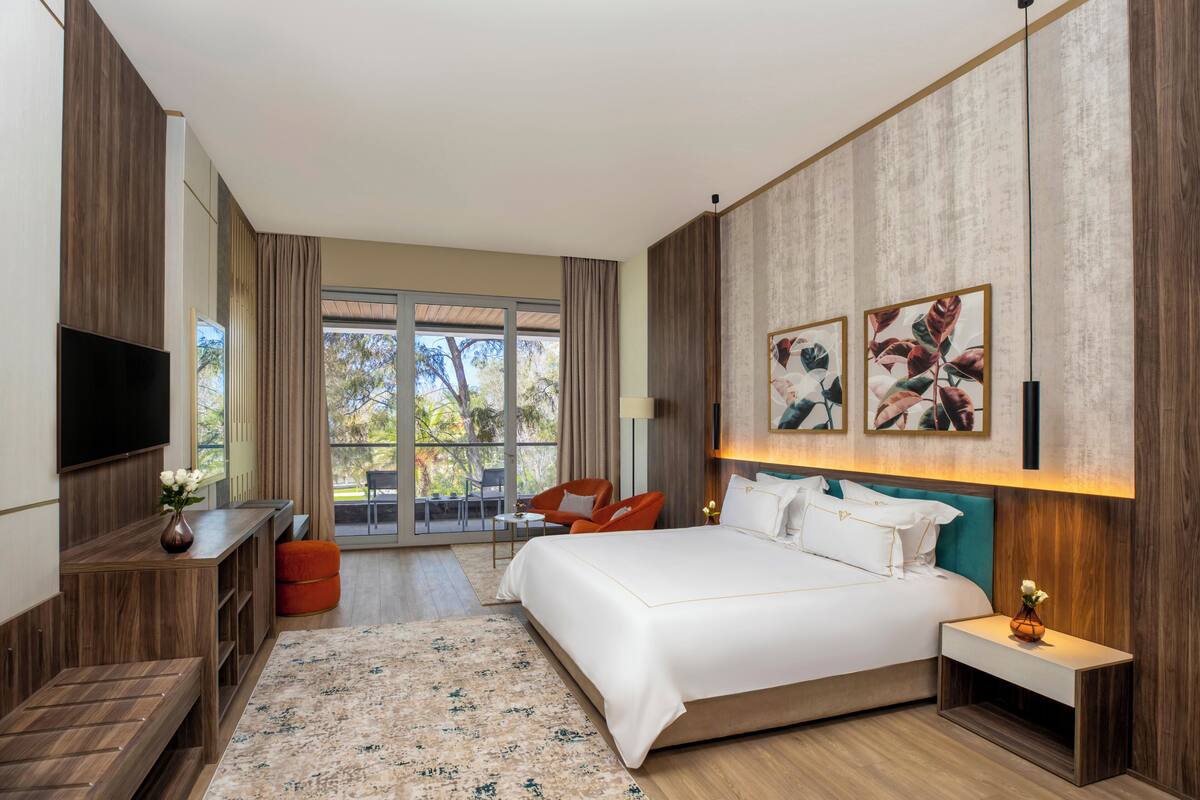 A modern hotel room with a large bed, a flat-screen TV, a desk, and a seating area with a view of the outdoors.