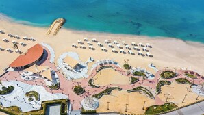 An aerial view of a beach resort with a large building, multiple beach umbrellas, and a well-maintained walkway. - Rixos Montaza Alexandria (Alexandria)