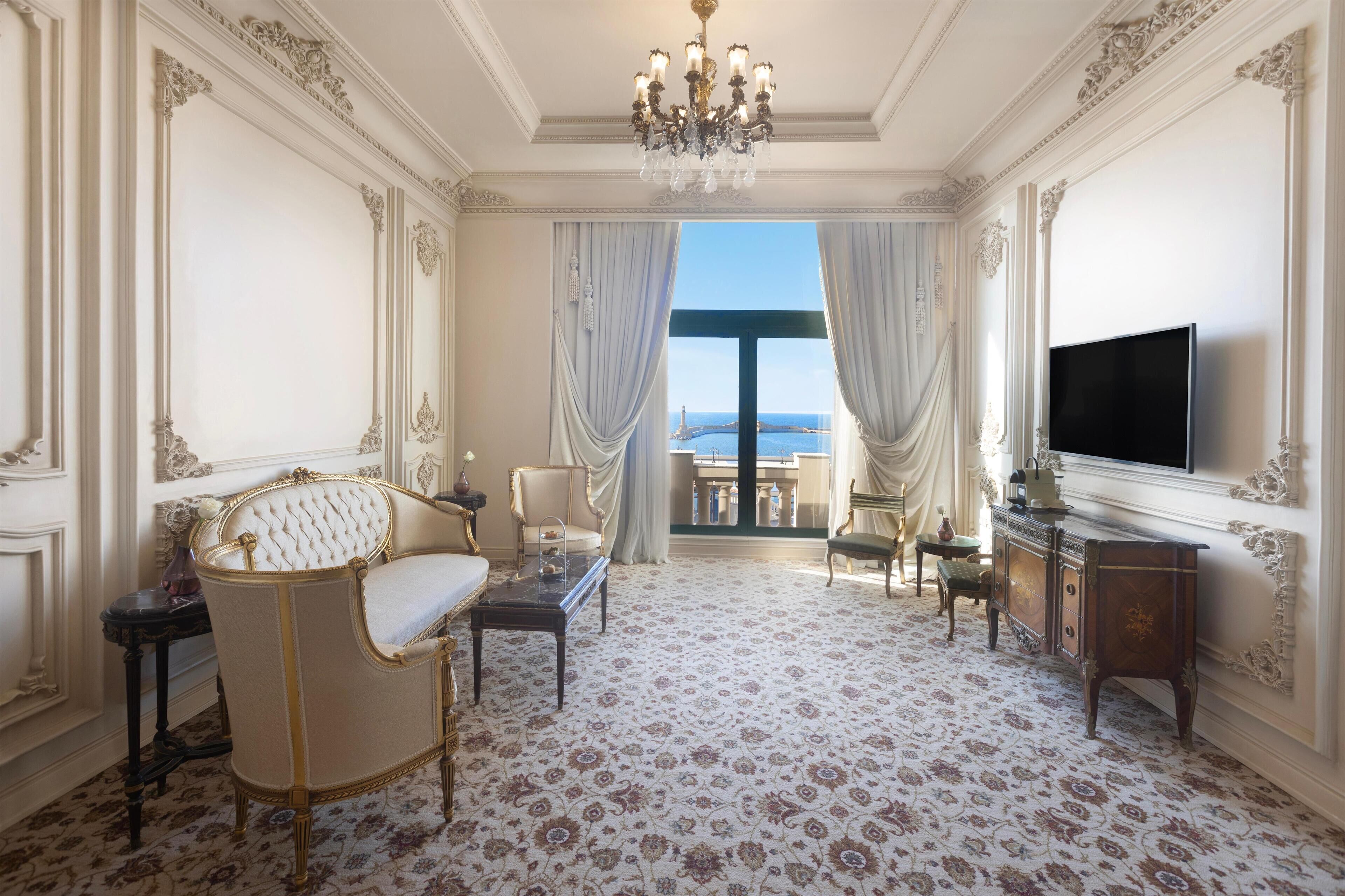 A luxurious room with ornate decor, a large chandelier, a sofa, a television, and a view of the sea through a large window.