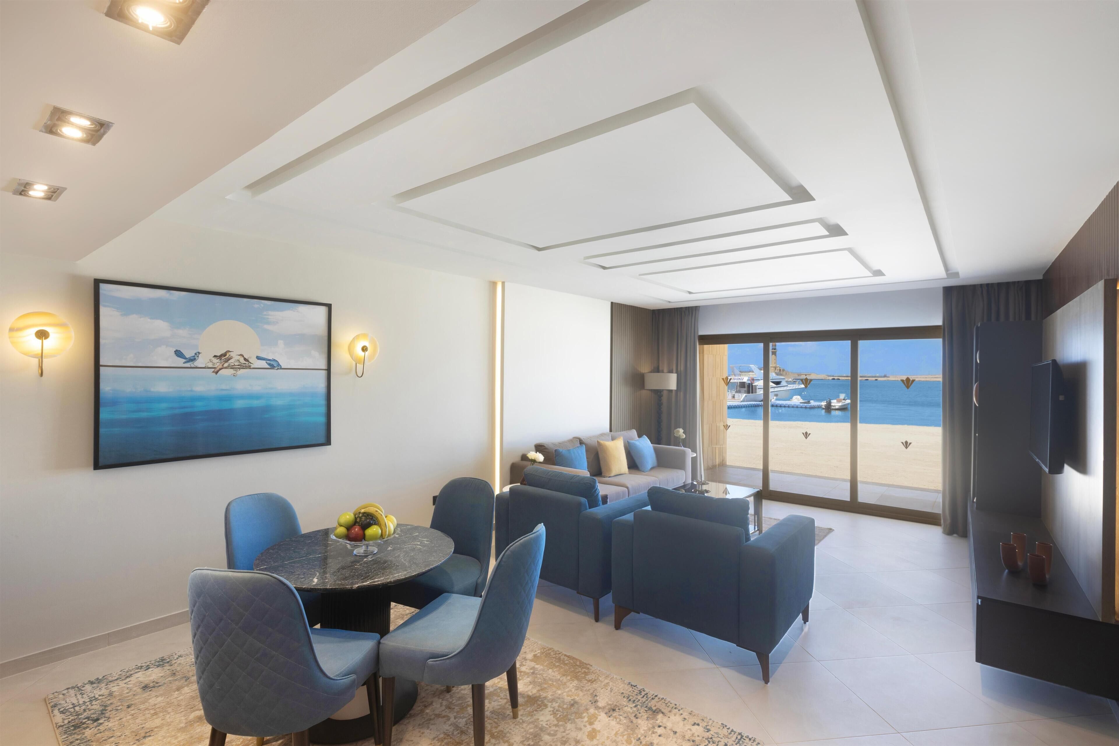 Uma sala de estar moderna com área de jantar, um conjunto de sofás e uma grande janela com vista para a praia e barcos.