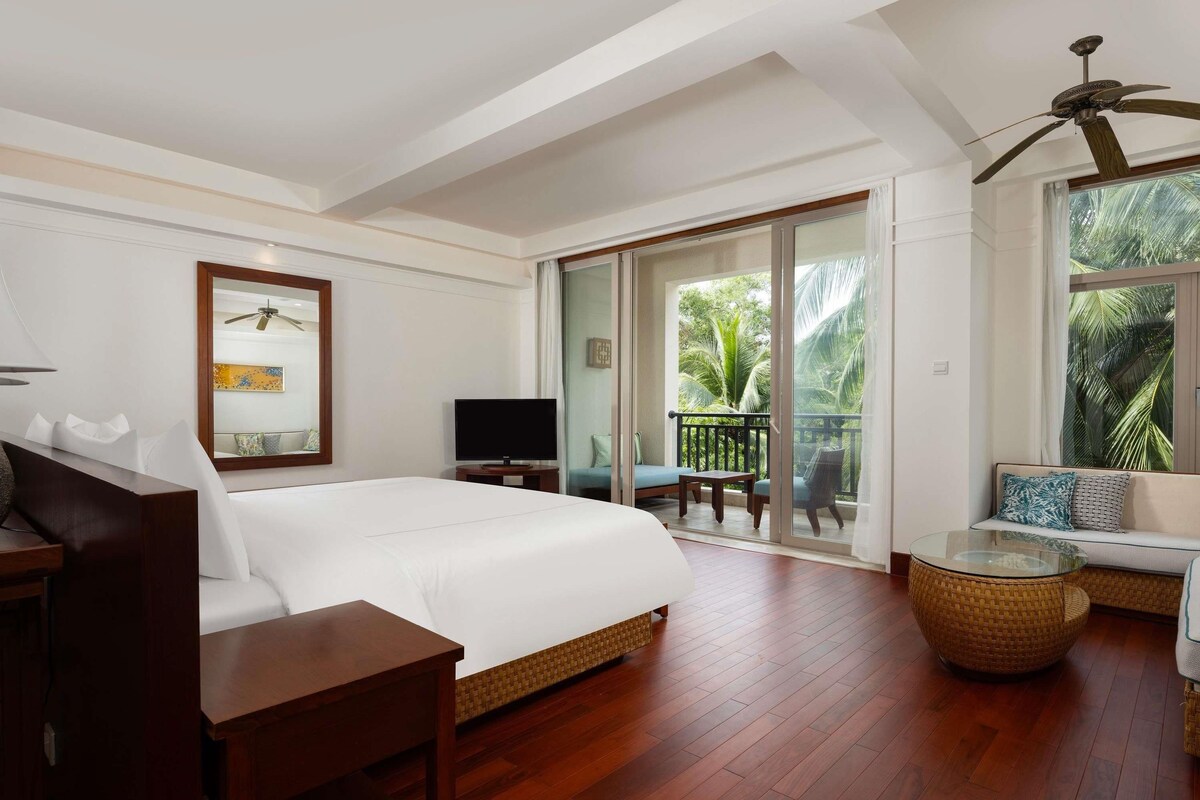 Een hotelkamer met een bed, een televisie, een balkon met uitzicht op palmbomen en een plafondventilator.