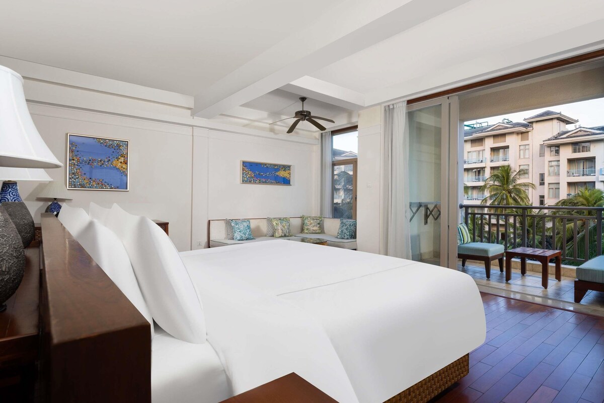 Een hotelkamer met een bed, een bureau, een stoel, een plafondventilator en een balkon met uitzicht op het gebouw.