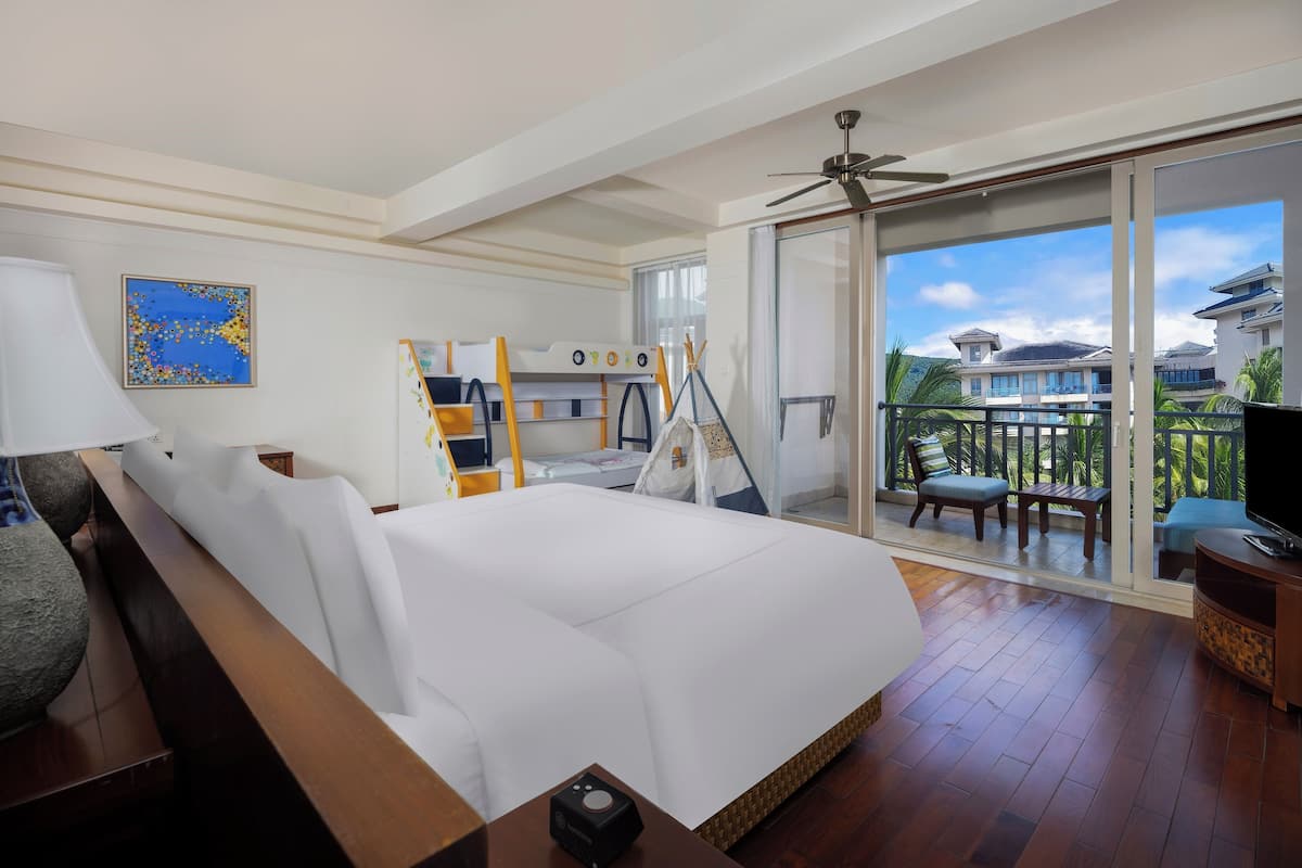 Een hotelkamer met een groot bed, een plafondventilator, een televisie en een balkon met uitzicht op andere gebouwen en palmbomen.