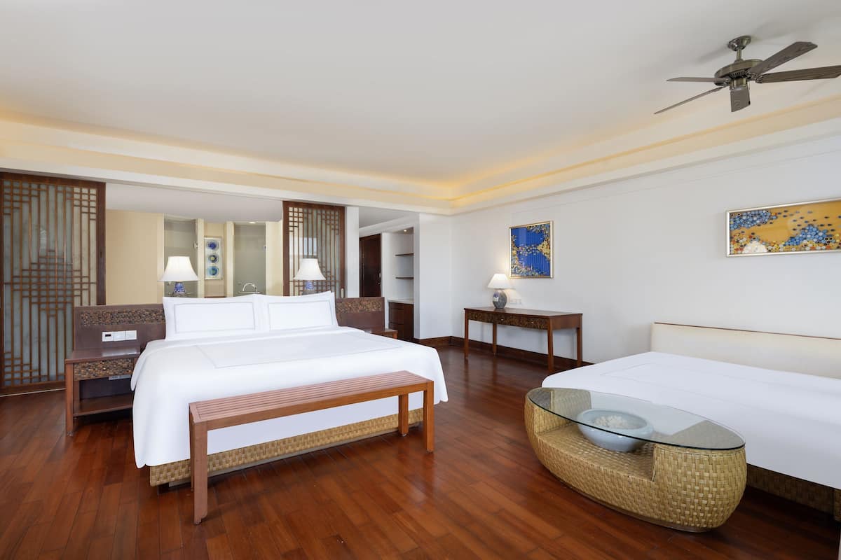 Hotelkamer met een groot bed, een glazen salontafel, nachtkastjes met lampen, een plafondventilator en een decoratief wandobject.