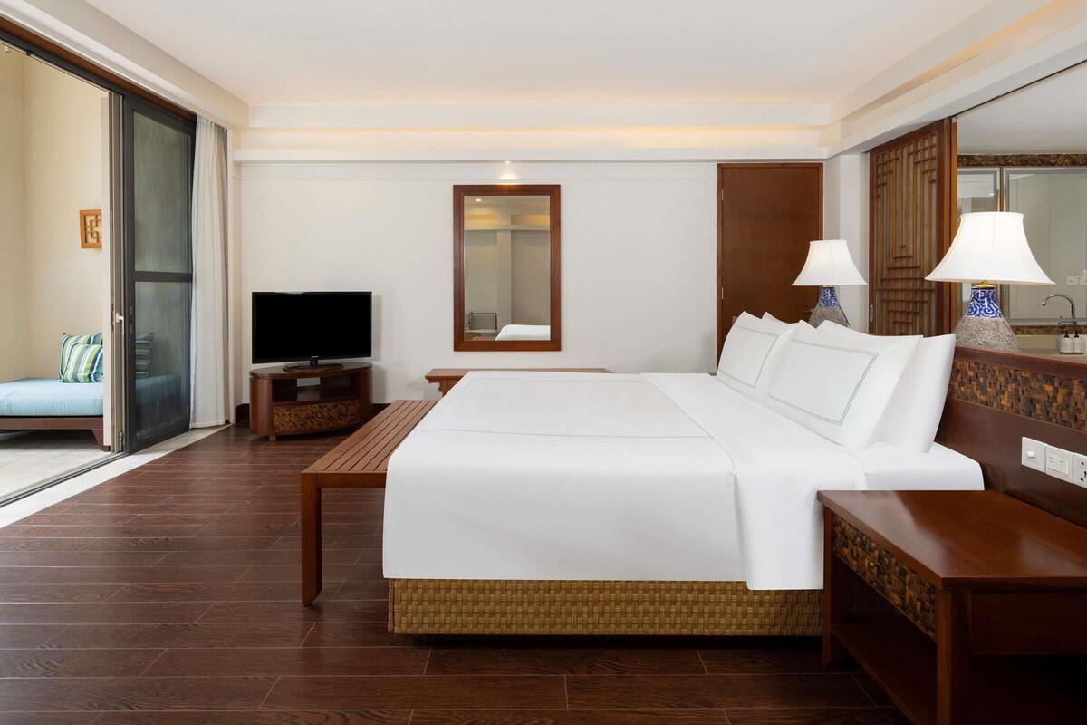 Een hotelkamer met een groot bed, een televisie, een houten nachtkastje, een rieten bijzettafeltje en een glazen schuifdeur die leidt naar een balkon.