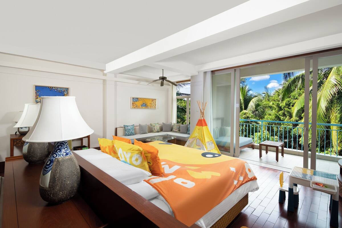 Een slaapkamer met een bed, kussens, een lamp, een bank en een balkon met uitzicht op groen.