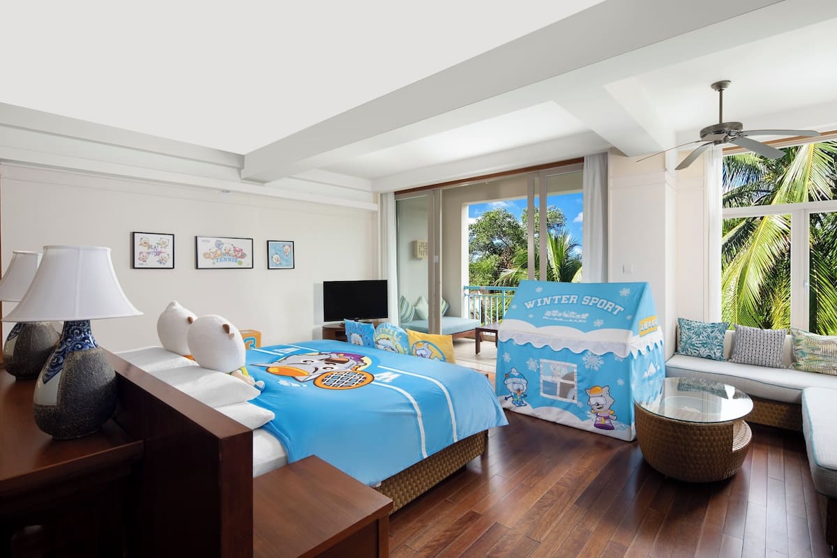 Een slaapkamer met een bed, een televisie, een bank, een raam met uitzicht op palmbomen en een plafondventilator.