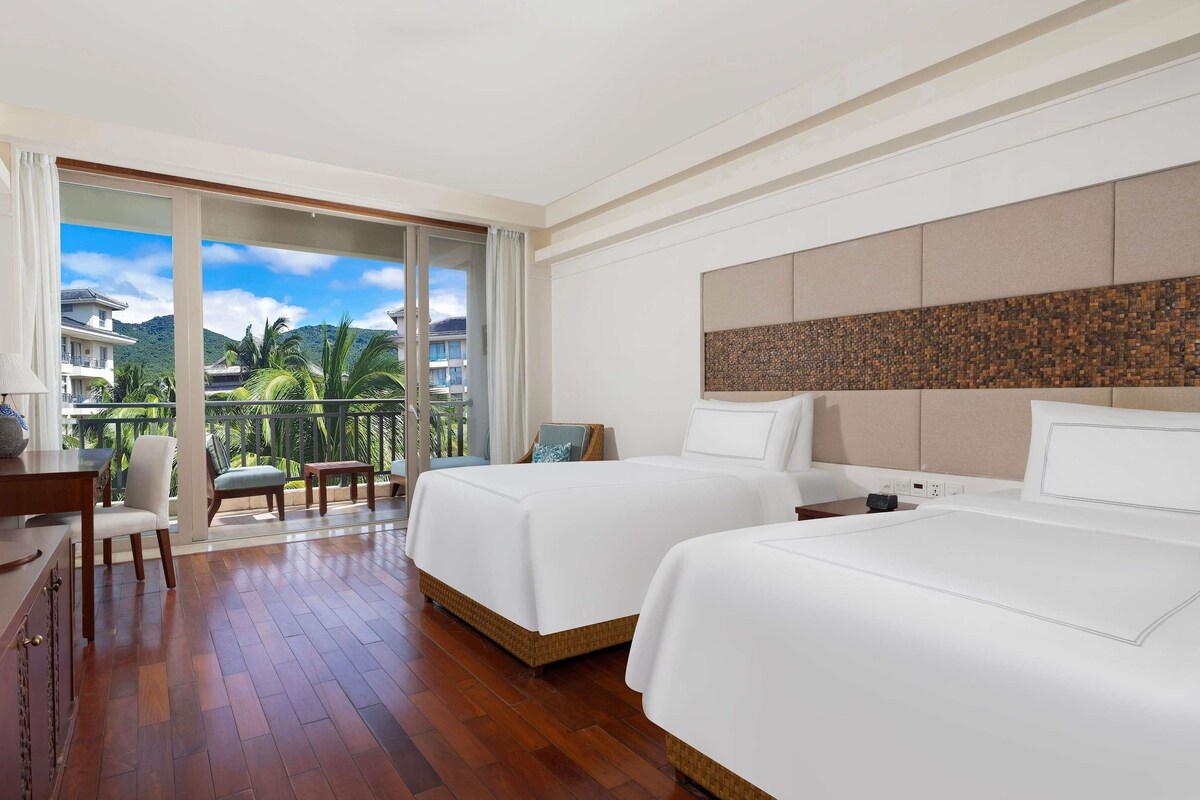 Een hotelkamer met twee bedden, een balkon met uitzicht op palmbomen en gebouwen, en een houten vloer.