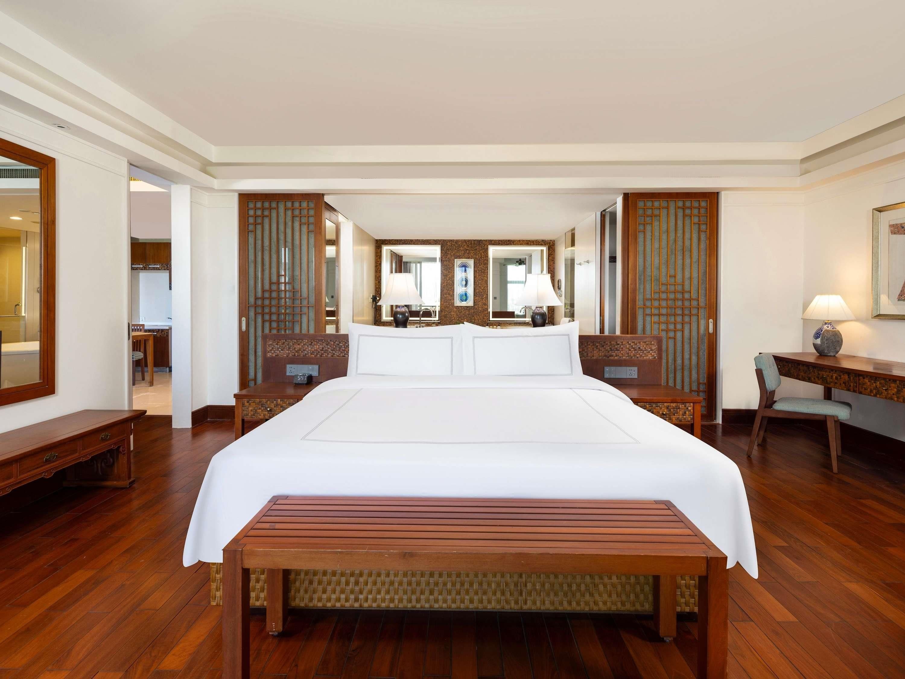 Una habitación de hotel con una cama grande, un banco de madera, un escritorio con una lámpara y un espejo.