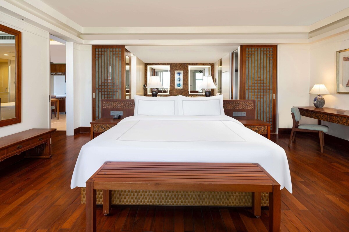 Een hotelkamer met een groot bed, een houten bankje, een bureau met lamp en een spiegel.