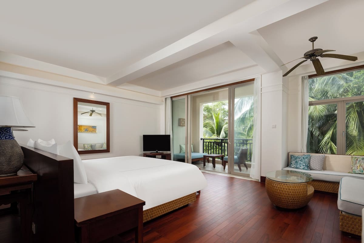 Een ruime slaapkamer met een groot bed, een televisie, een balkon met uitzicht op palmbomen en een plafondventilator.