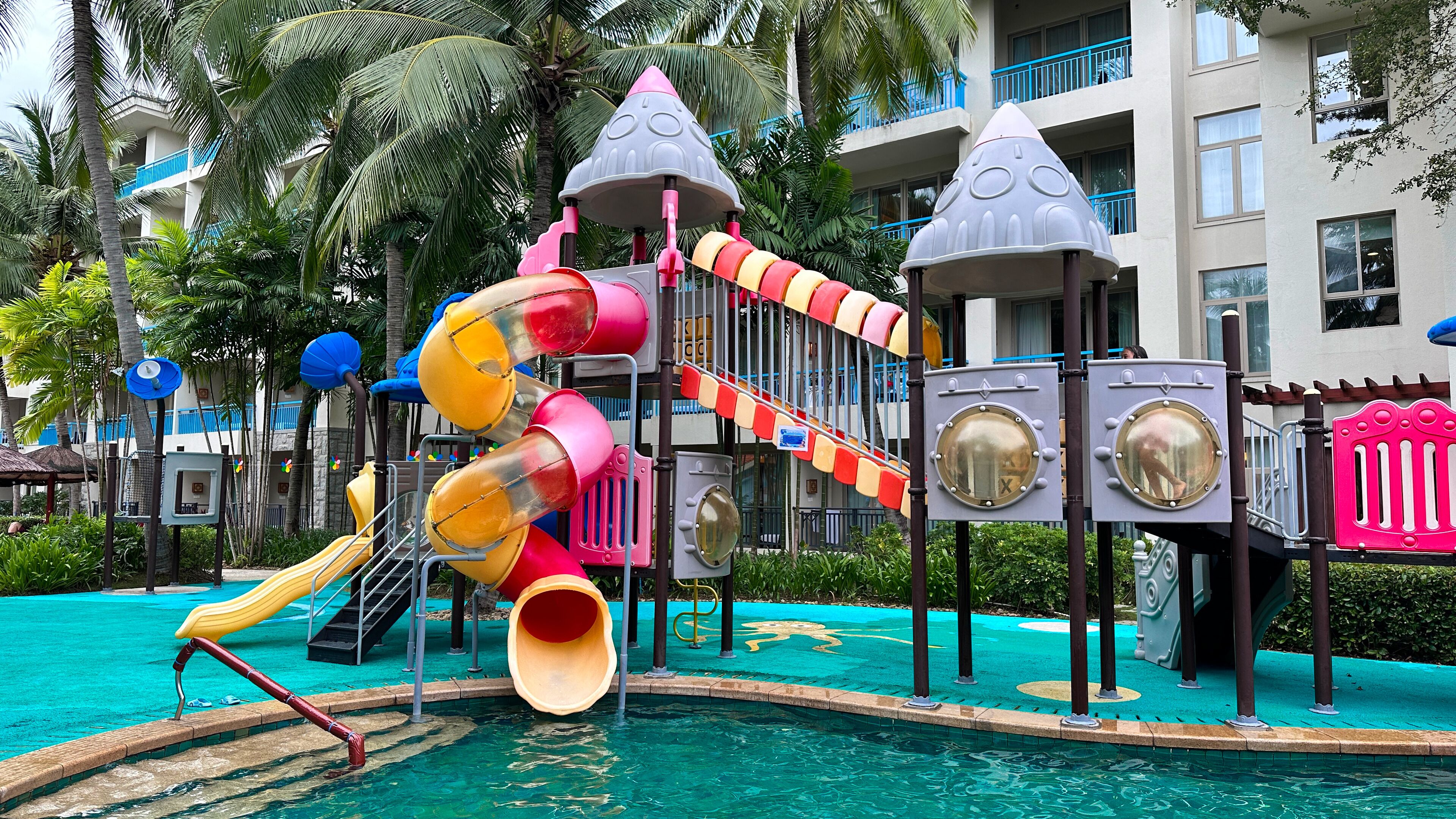 A children's play area with slides and climbing structures, surrounded by a pool and palm trees.