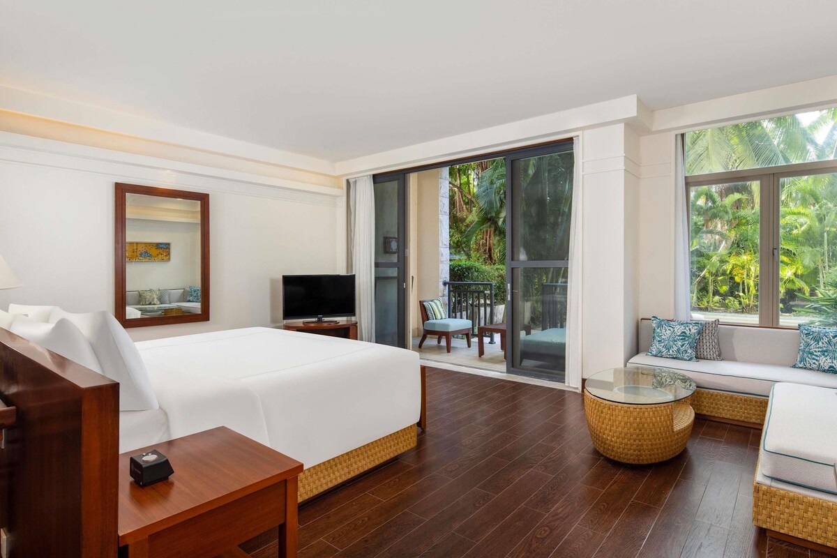 Hotelkamer met een bed, een televisie, een bank, een glazen tafel en een balkon met uitzicht op groen.