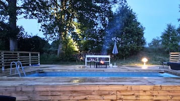 A wooden deck with a pool, a table with chairs, and a fire pit.