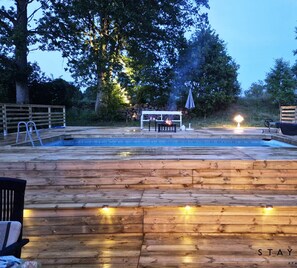 Une terrasse en bois avec une piscine, une table entourée de chaises et un foyer extérieur.