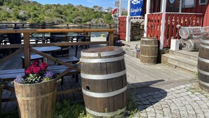A wooden deck with tables and barrels, a red building with a sign, and a view of a hill. - Large villa with sea view in Rönnäng | SE09078 (Rönnäng)