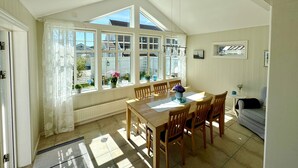 A dining area with a wooden table and chairs, large windows with white curtains, and a view of the outside.