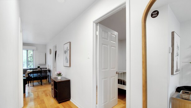 Un pasillo con piso de madera, paredes blancas y una puerta que da a una habitación. Se ve un comedor con una mesa y sillas al fondo.