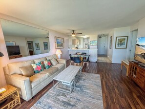 A living room with a sofa, coffee table, and dining area. There is a kitchen in the background with a dining table and chairs.