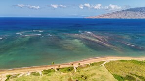 An aerial view of a coastal area with clear blue waters, a sandy beach, and a distant hill with wind turbines.