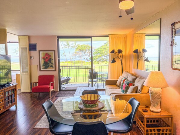 A living room with a glass table, chairs, a sofa, and a balcony with a view of the ocean.